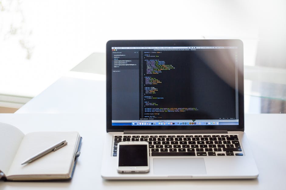 A silver laptop on a white desk displaying lines of colorful code on its screen, positioned centrally with a black smartphone placed in front of it. To the left of the laptop, an open white notebook w